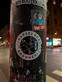 Street sticker The sticker is circular with a white background and black design elements. In the center, there are two horizontal wavy lines, symbolizing water or waves. Above these waves, a delicate botanical branch with leaves and small berries or buds is depicted. Encircling these elements, the text 'BARSEBÄCKS' is written along the top curve, and 'BRÄNNERI' along the bottom curve, both in a clean, sans-serif font. The overall aesthetic is minimalist and elegant, suggesting a brand identity for a distillery.