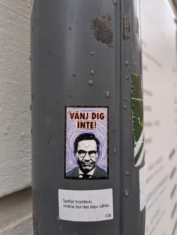 Street sticker A rectangular sticker featuring a stylized, black and white portrait of a man, identifiable as Stefan Löfven, from the chest up. He wears glasses, a suit jacket, and a striped shirt with a tie, and has a serious expression. Behind his head, a background of concentric, wavy purple lines on a white base creates a dynamic effect. Above his head, the text 'VÄNJ DIG INTE!' is prominently displayed in bold orange capital letters with a black outline. The sticker is framed by a thin black border, an orange border, and a thicker black border adorned with small white dots. It is affixed to a wet, dark grey pole.