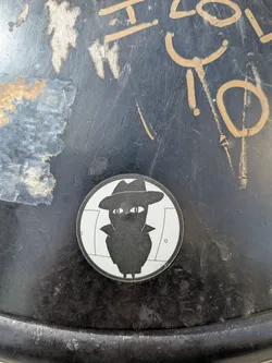 A round sticker showing a black silhouette of a cat-like figure wearing a fedora and trench coat. The figure has white, slit-pupil eyes, set against a minimalist white background with simple line-drawn doors.