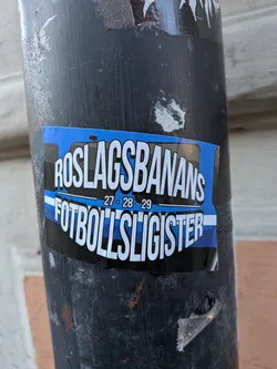 Street sticker A rectangular sticker on a dark pole featuring blue, black, and white horizontal stripes. Bold white text reads 'ROSLAGSBANANS' on top and 'FOTBOLLSLIGISTER' on the bottom, with '27 - 28 - 29' in the center.