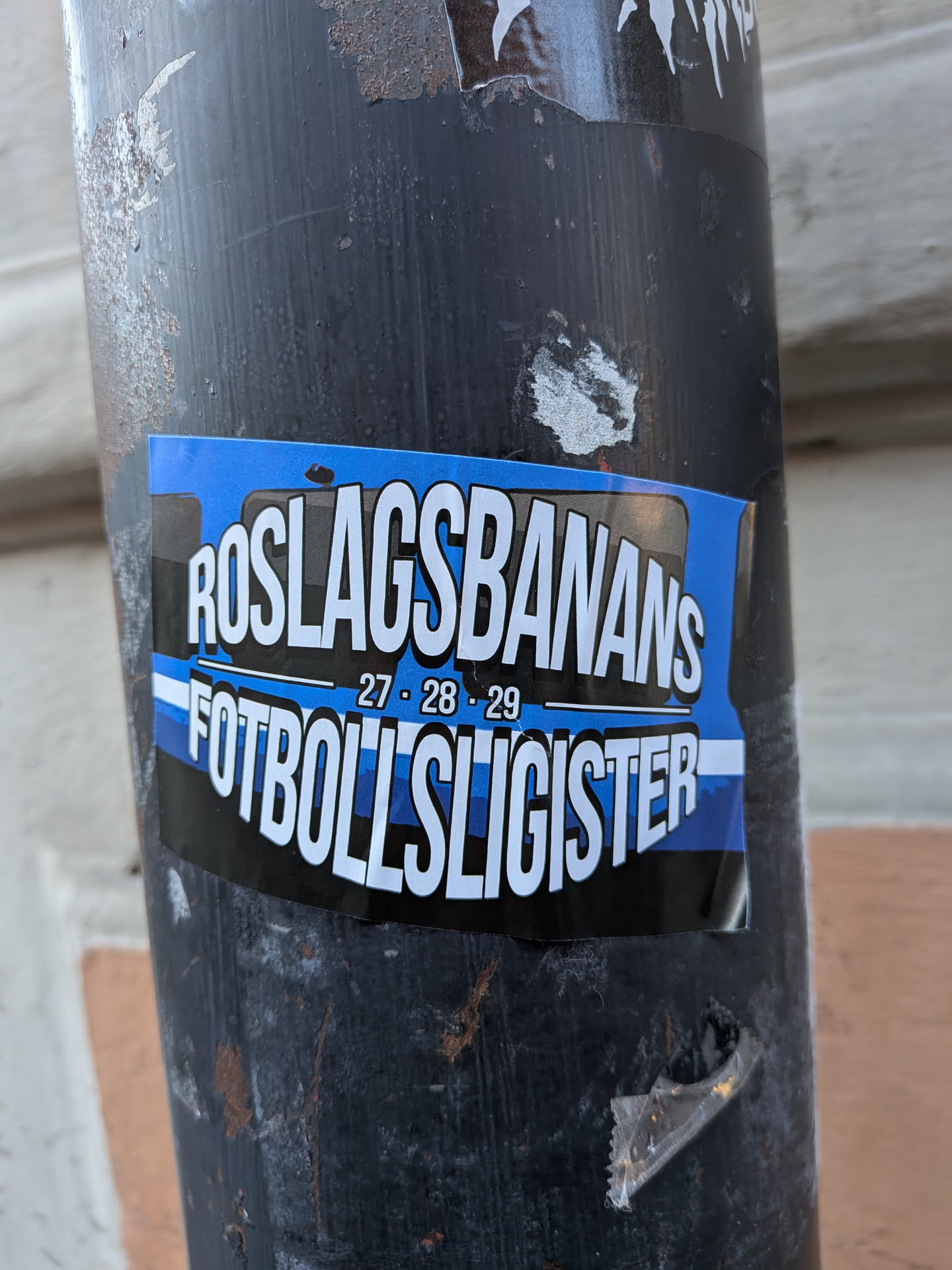 A rectangular sticker on a dark pole featuring blue, black, and white horizontal stripes. Bold white text reads 'ROSLAGSBANANS' on top and 'FOTBOLLSLIGISTER' on the bottom, with '27 - 28 - 29' in the center.