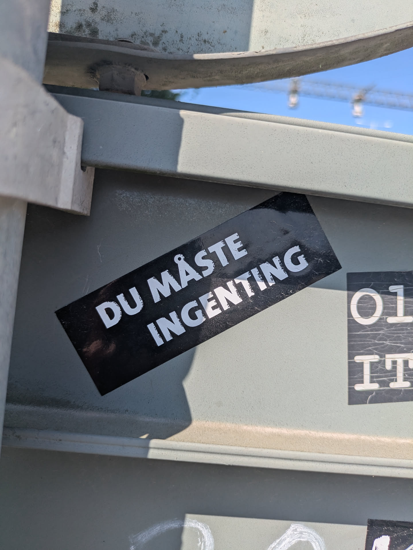 A black sticker with white text reading "DU MÅSTE INGENTING", placed on a grey metal structure. The text translates to 'YOU MUST NOTHING'. Another partial sticker is on the right.