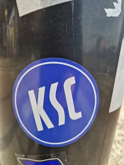 Street sticker A round blue sticker with the letters KSC in a stylized sans-serif font. The sticker is affixed to a dark-colored pole.
