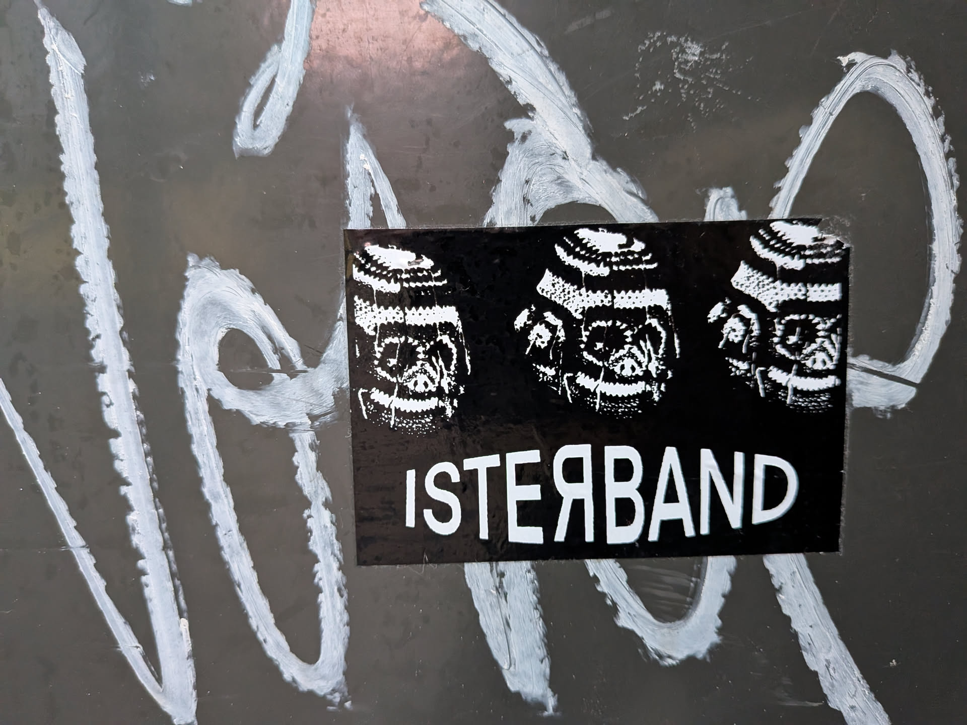 Street sticker The sticker is rectangular with a black background. It features three monochrome, stylized skull illustrations arranged horizontally across the top half. The skulls have intricate patterns within their eye sockets and teeth, rendered in white. Below the skulls, in bold white sans-serif lettering, is the text 'ISTERBAND'. The sticker appears to be applied to a dark, possibly metallic surface that has been marked with white graffiti.