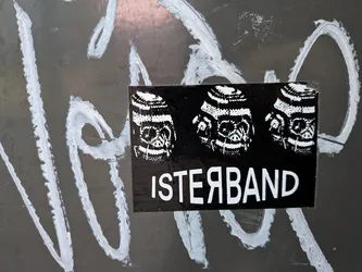 Street sticker The sticker is rectangular with a black background. It features three monochrome, stylized skull illustrations arranged horizontally across the top half. The skulls have intricate patterns within their eye sockets and teeth, rendered in white. Below the skulls, in bold white sans-serif lettering, is the text 'ISTERBAND'. The sticker appears to be applied to a dark, possibly metallic surface that has been marked with white graffiti.