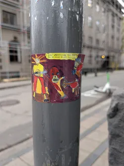 Street sticker The sticker features a colorful, hand-drawn illustration with a dark maroon background. In the center, a stylized figure with a yellow body, red pants, and a mask-like face with orange rays emanating from its head is depicted playing a banjo. To the right, a red, monstrous creature with sharp teeth and a distorted face lunges forward, with a smaller, brown, deer-like creature in front of it. The overall style is crude and cartoonish, with bold outlines and vibrant colors. A yellow banner runs across the top of the sticker with text.