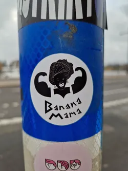 A sticker featuring a strong woman with biceps, wearing a head wrap. The text 'Banana Mama' is written below the illustration. The sticker is predominantly black and white with a circular design, and it's pasted on a blue surface.