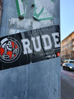 A black and white sticker with the word "RUDE" prominently displayed.  It features a circular logo with a skull wearing a hat.  Smaller text indicates an address and website. The style is reminiscent of punk or rockabilly aesthetics. 