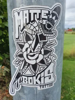 Black and white sticker with a tattoo-style design. It features a rose, a dagger, and stylized lettering. The text 'Matte Bokis Tattoo' is prominently displayed.
