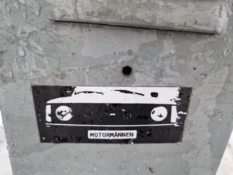 Street sticker A black and white sticker featuring the front of a classic car. The word "MOTORMANNEN" is visible at the bottom. The sticker is affixed to a weathered grey metal surface.