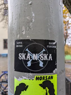 A black and white sticker with the text "Ska N Ska" in the center. Two trumpets are crossed behind the text. At the top of the sticker, it says "Love Music" on the left and "Hate Fascism" on the right. The sticker is attached to a pole.