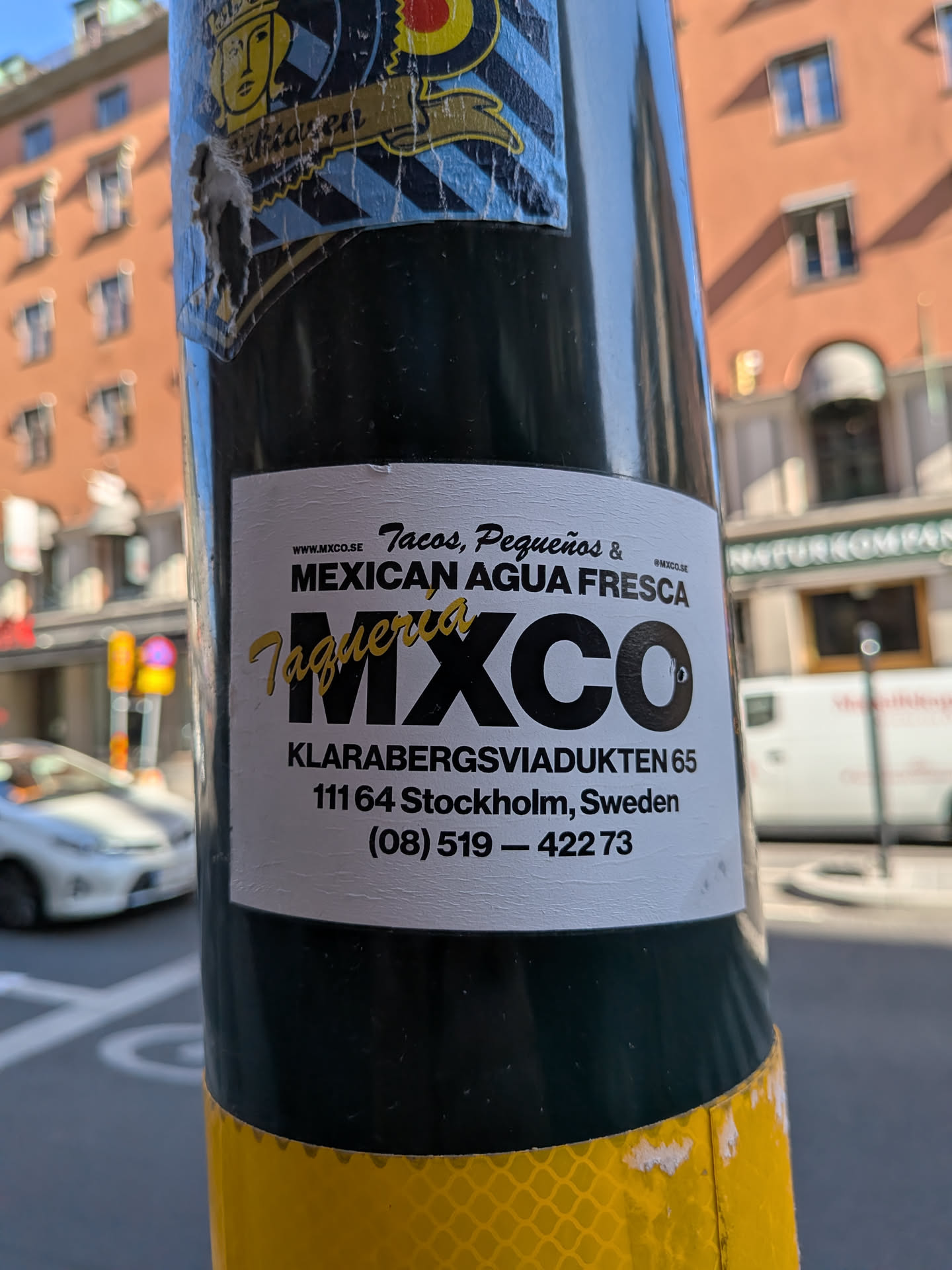 A white rectangular sticker featuring black and yellow typography. It advertises Taqueria MXCO, displaying their menu items, Stockholm address, phone number, and website.