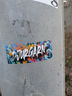 Street sticker The sticker has a blue, orange, white and black background, with the word "Djurgarn" written in white. The sticker is on a gray pole that is a bit dirty. There is also another sticker above it.