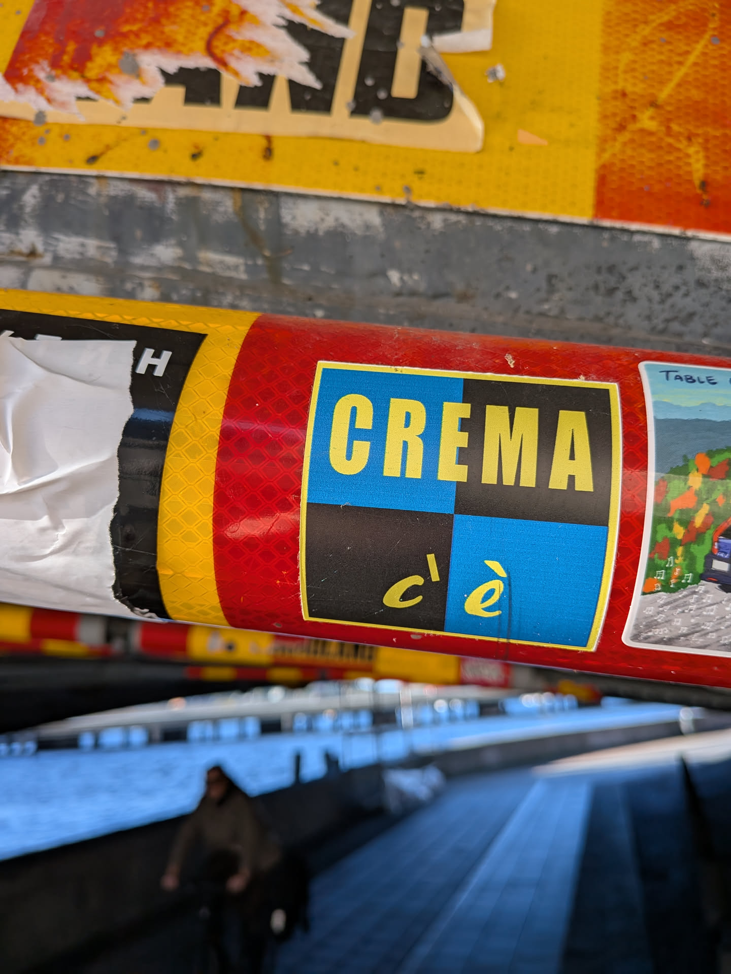 The sticker is predominantly red with a yellow border. In the center, a blue and black checkered square contains the word 'CREMA' in large, bold yellow sans-serif letters. Below this, in smaller yellow script, are the words 'c'è'. To the right of this central design is a small, colorful illustration depicting a landscape with trees, a car, and musical notes. The sticker appears to be applied to a cylindrical object, possibly a tube or a can, and shows signs of wear and tear, with some peeling white material on the left side.
