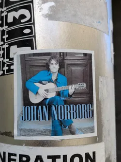 This sticker showcases musician Johan Norborg with his guitar. He is dressed in a blue outfit and is sitting in front of a doorway. The sticker appears to be placed on a pole, adding a touch of artistic expression to the urban landscape.