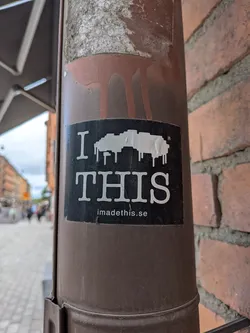 Street sticker The sticker is rectangular with a black background. In the center, a prominent white pixelated shape appears to be melting or dripping, resembling an abstract cloud or a distorted, dripping object. Above this shape, in a large white serif font, is the letter "I", and below it, also in large white serif font, is the word "THIS". A small registered trademark symbol (®) is visible to the upper right of the pixelated shape. At the very bottom of the sticker, in smaller white sans-serif font, is the website address "imadethis.se". The sticker shows some minor wear and tear, consistent with being affixed outdoors.