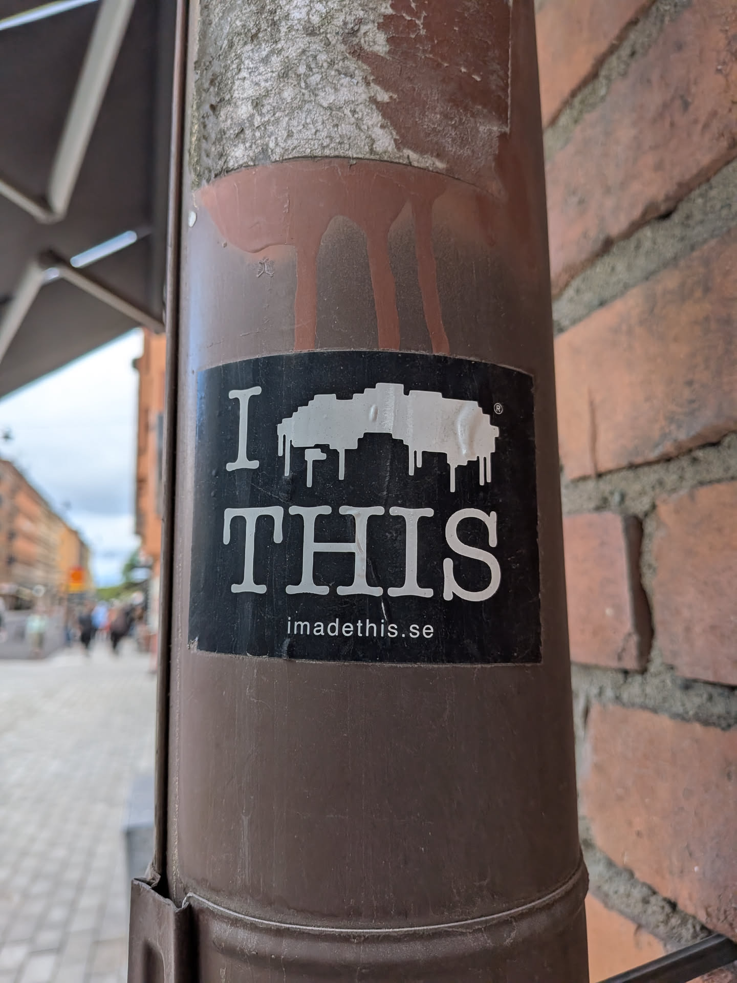 The sticker is rectangular with a black background. In the center, a prominent white pixelated shape appears to be melting or dripping, resembling an abstract cloud or a distorted, dripping object. Above this shape, in a large white serif font, is the letter "I", and below it, also in large white serif font, is the word "THIS". A small registered trademark symbol (®) is visible to the upper right of the pixelated shape. At the very bottom of the sticker, in smaller white sans-serif font, is the website address "imadethis.se". The sticker shows some minor wear and tear, consistent with being affixed outdoors.