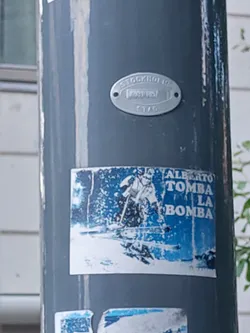 Street sticker A sticker featuring Alberto Tomba, an Italian former World Cup alpine ski racer, with text that says 'ALBERTO TOMBA LA BOMBA'. The sticker is faded and worn, attached to a grey pole.