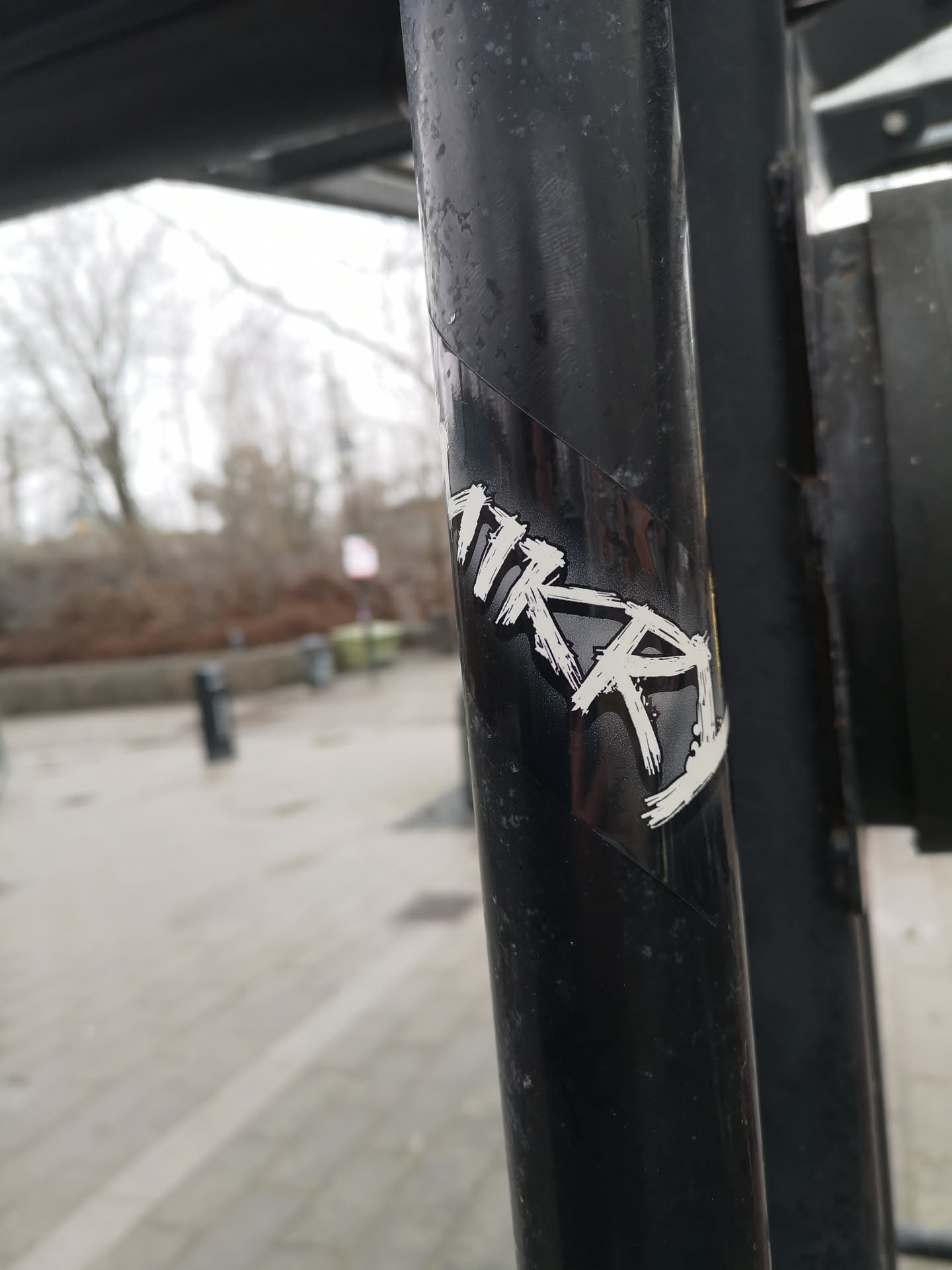 The sticker is rectangular, predominantly black with white text, and is wrapped around a dark, possibly wet, metal pole. The text, which appears to read 'MKRA', is rendered in a distressed, brushstroke or graffiti-style font, giving it a hand-painted or spray-painted aesthetic. The white letters stand out sharply against the glossy black background of the sticker. The sticker shows some minor wear and tear, including small scuffs and possibly water marks, consistent with outdoor exposure.