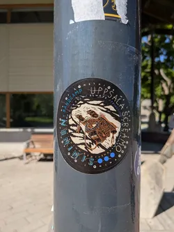 Street sticker A circular sticker featuring a central illustration of a stylized skull or skeletal face intertwined with what appears to be an engine block or mechanical components, all set against a backdrop of swirling, wave-like patterns in a light beige or off-white color. The mechanical elements are rendered in shades of brown and orange, with intricate line work. Surrounding this central image, text is arranged in arcs. The outer ring features 'SOMETHING WE LOST' at the top and 'SOMETIMES WE WIN' at the bottom, in a light blue font. The inner ring includes 'BANDCAMP' and 'UPPSALA SCREAMO' at the top, and 'NININ' repeated with blue dots at the bottom. Above the central illustration, 'ALWAYS STRUGGLING' is written. The overall color scheme is dark (black background) with light beige, brown, orange, and light blue accents.