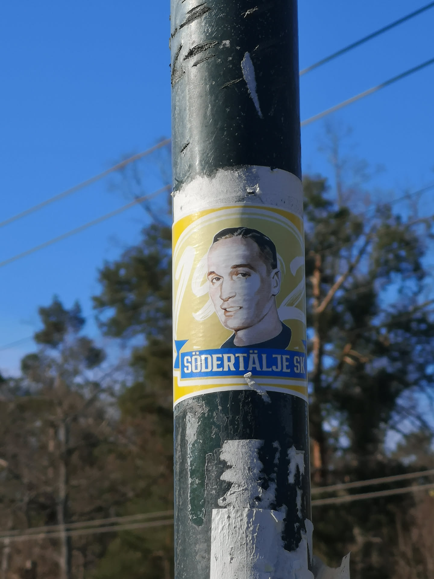 A rectangular sticker featuring a stylized portrait of a man with short, dark hair and a subtle smile, looking slightly to the right. He is depicted from the chest up, wearing a dark blue shirt, against a bright yellow background with subtle white swirling lines. Below the portrait, a blue banner with a white border displays the text 'SÖDERTÄLJE SK' in white capital letters. The sticker is affixed to a dark, weathered pole, showing some wear and tear around its edges.