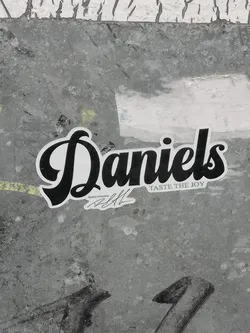 The sticker prominently displays the word 'Daniels' in a bold, flowing black script font with a clean white fill and a thin black outline. Below 'Daniels' and slightly to the right, the slogan 'TASTE THE JOY' is written in a smaller, sans-serif font. Further below and to the left, in an even smaller font, are the words 'Quality Guarantee' followed by a stylized black signature. The sticker has a distinct white border around all its elements, making it stand out against the textured, mottled gray background it's adhered to.