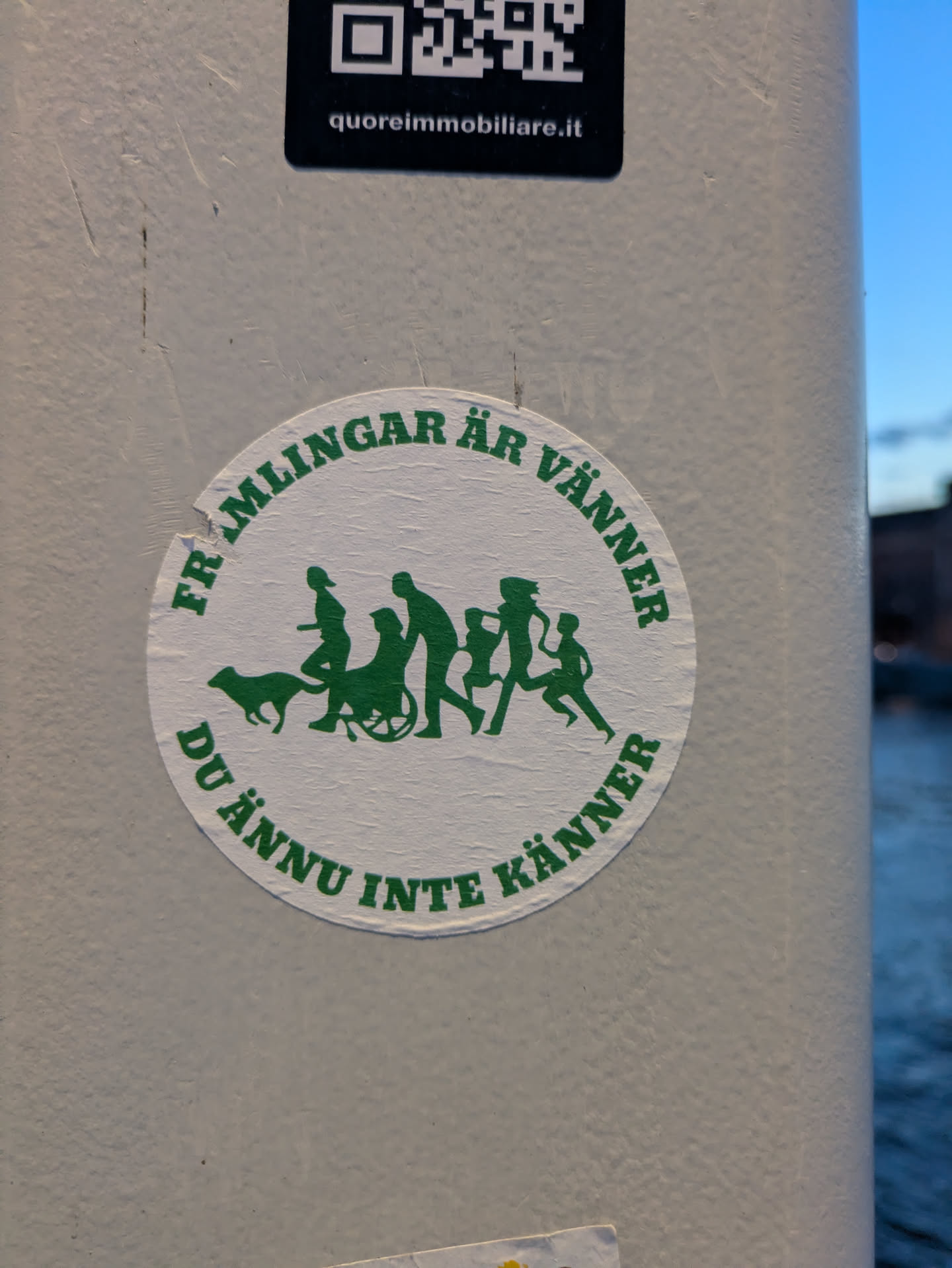 A circular sticker with a white background and dark green illustrations and text. It features a central horizontal line of stylized silhouettes depicting a diverse group of figures walking from left to right: a person walking a dog, a person in a wheelchair, an adult, and two children. The text "FRÄMLINGAR ÄR VÄNNER DU ÄNNU INTE KÄNNER" (Swedish for "Strangers are friends you haven't met yet") curves around the top and bottom edges of the circle. The sticker shows some signs of wear and tear, including minor creases and scuffs, indicating it is an actual physical sticker affixed to a textured surface.