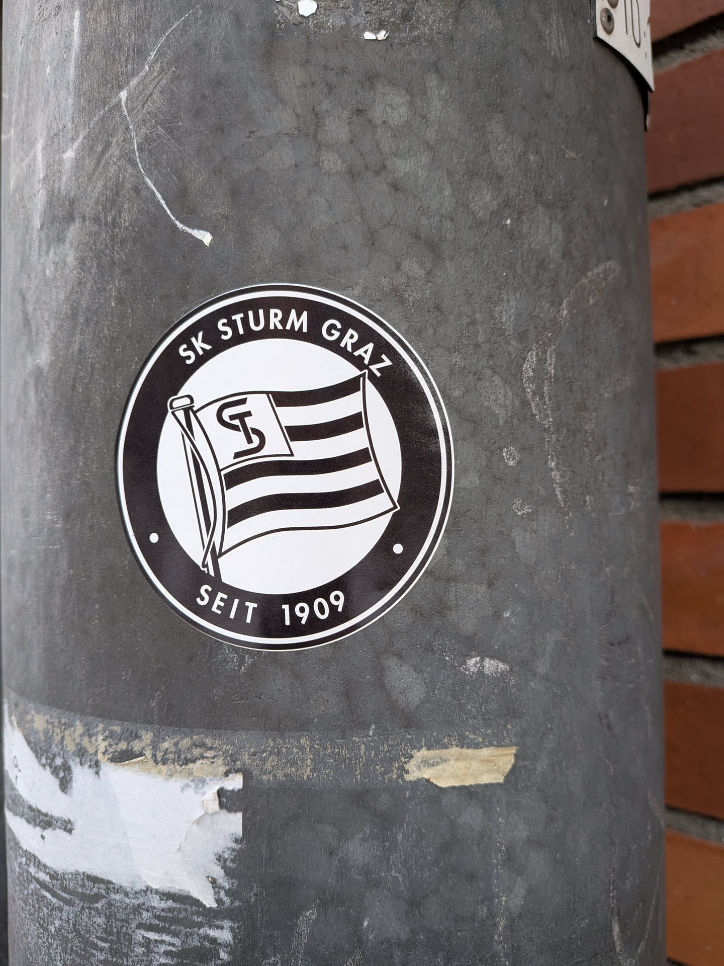 Street sticker A circular, monochrome sticker featuring the logo of SK Sturm Graz. The design is primarily black and white, with a thick black outer ring containing the text 'SK STURM GRAZ' at the top and 'SEIT 1909' at the bottom, separated by small white dots. The center of the sticker displays a stylized flag, waving from a flagpole, with horizontal stripes. A prominent, intertwined monogram of 'S' and 'T' is placed in the upper left quadrant of the flag. The overall appearance is clean and classic, emphasizing the club's identity and heritage.