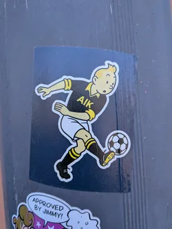 A rectangular sticker featuring a cartoon character, strongly resembling Tintin, with blonde hair and fair skin, in the act of kicking a black and white soccer ball. He is dressed in a black short-sleeved jersey with yellow trim on the collar and sleeves, prominently displaying the letters "AIK" in yellow on the chest. He wears white shorts, black socks with two yellow stripes, and black soccer cleats with yellow studs. The character is outlined in white against a dark blue background.