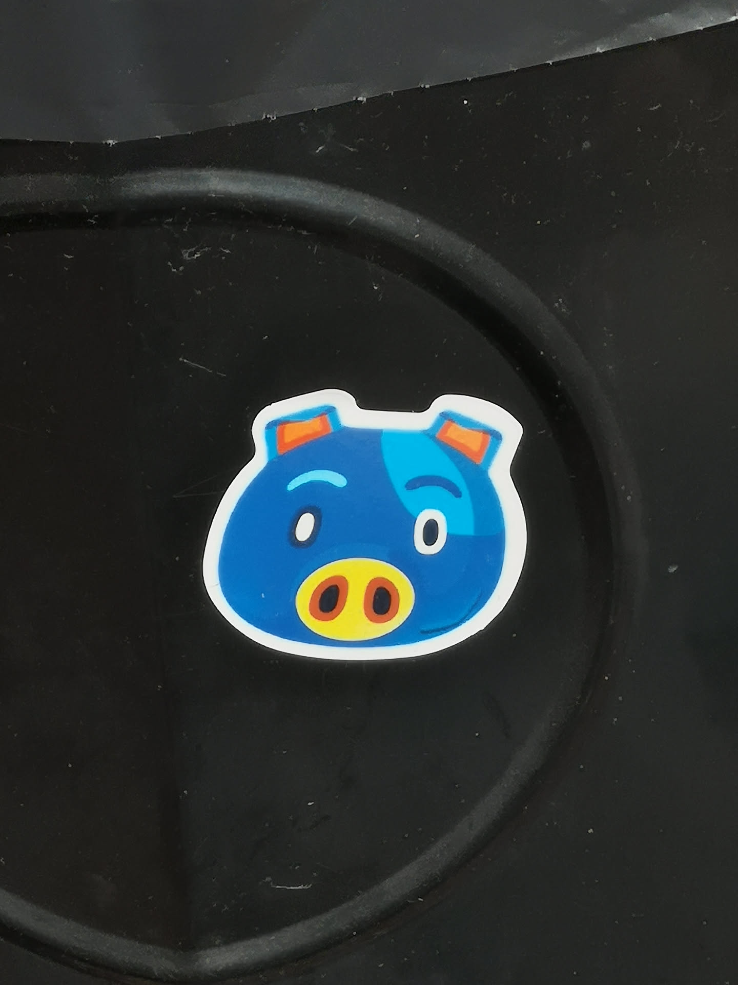 The sticker features a stylized, cartoon-like pig's head with a predominantly vibrant blue color. It has a rounded, irregular shape with small, orange-tipped ears at the top. The face includes a prominent yellow snout with two dark brown oval nostrils. Its eyes are white ovals with small black pupils, one slightly larger than the other, giving it a playful or slightly surprised expression. A lighter blue patch is visible on the right side of its head, and simple blue curved lines serve as eyebrows. The entire design is outlined with a clean white border, making it stand out against the dark background.