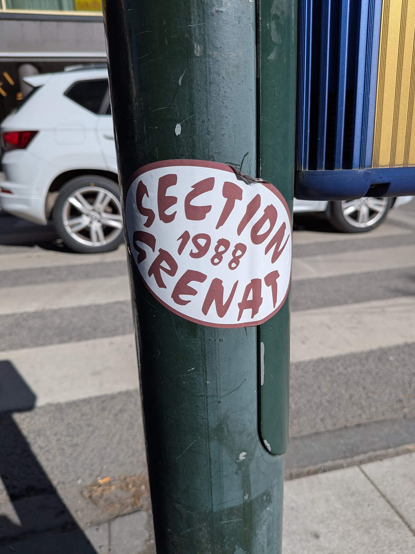 An oval-shaped sticker featuring a white background with a thin maroon border. The text 'SECTION 1988 GRENAT' is printed in a bold, somewhat distressed or hand-drawn style font, colored maroon. 'SECTION' is positioned at the top, '1988' is centered below it, and 'GRENAT' is at the bottom. The sticker shows minor wear, including a small tear at its top edge, and is affixed to a dark green metal pole.