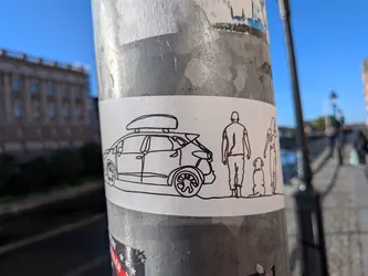 Street sticker The sticker is a minimalist, black and white outline drawing on a white rectangular background. It depicts a car with a roof box, followed by a silhouette of a person, a dog sitting, and another person standing. The style is simple and linear, with no shading or color fill.