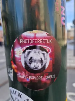 A round sticker featuring a ferret's face against a blurry background of neon lights.  The text "PHOTOFERRETUK" is at the top, and "TRAVEL - EXPLORE - CREATE" is at the bottom. 
