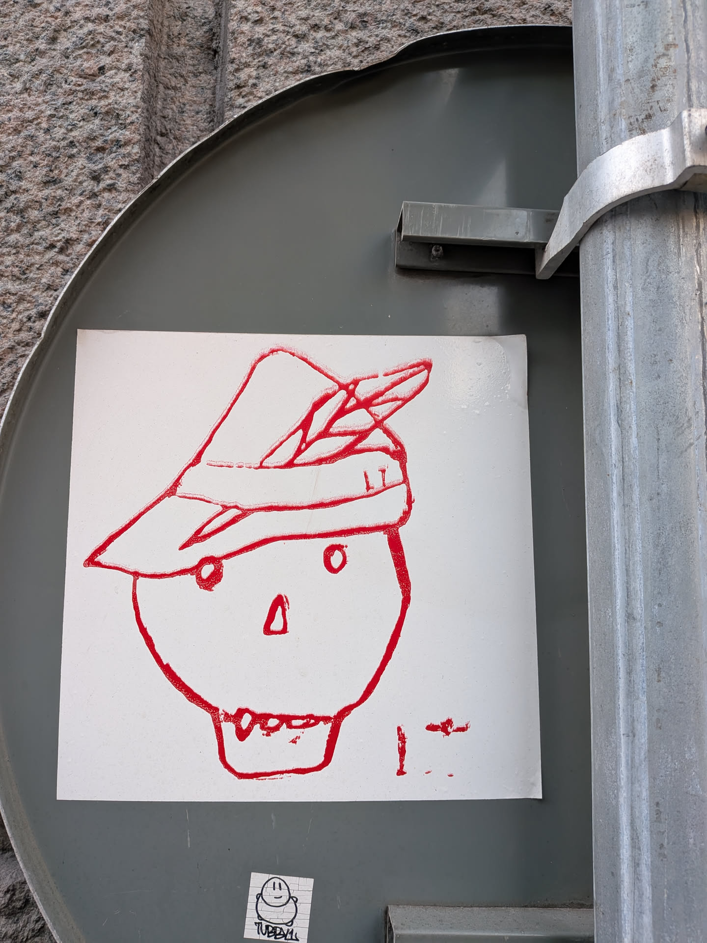 Street sticker The sticker features a simple, hand-drawn red outline of a skull wearing a hat adorned with a feather. The skull has two small circles for eyes, a triangular nose, and a mouth with visible teeth. The hat is a traditional Bavarian-style cap. To the right of the skull, there are some red marks that appear to be abstract scribbles or a signature. Below the main drawing, there is a smaller, separate sticker with a black and white drawing of a smiling character and the text 'TUBBYLL'.