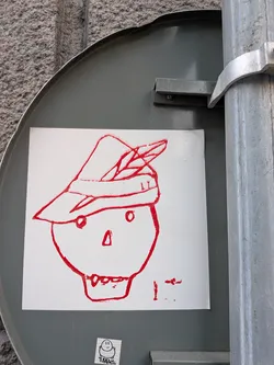 Street sticker The sticker features a simple, hand-drawn red outline of a skull wearing a hat adorned with a feather. The skull has two small circles for eyes, a triangular nose, and a mouth with visible teeth. The hat is a traditional Bavarian-style cap. To the right of the skull, there are some red marks that appear to be abstract scribbles or a signature. Below the main drawing, there is a smaller, separate sticker with a black and white drawing of a smiling character and the text 'TUBBYLL'.