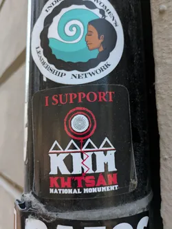 Street sticker The sticker is black with red and white text and graphics. At the top, it reads 'I SUPPORT' in red capital letters. Below this is a graphic with concentric red and white circles, resembling a target or a stylized vortex. Underneath this symbol are four white triangles, connected at their bases, forming a stylized mountain or pyramid shape. The letters 'KWT'SAN' are prominently displayed in white, bold, block letters with a jagged red line cutting through the 'M'. Below this, in smaller red capital letters, it says 'NATIONAL MONUMENT'. The sticker appears to be slightly worn and has some dust or debris on its surface.