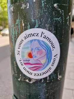 A round sticker with a drawing of a woman's face and a sunset in the background. The text around the image says "If you love love, you'll love my art." in French.