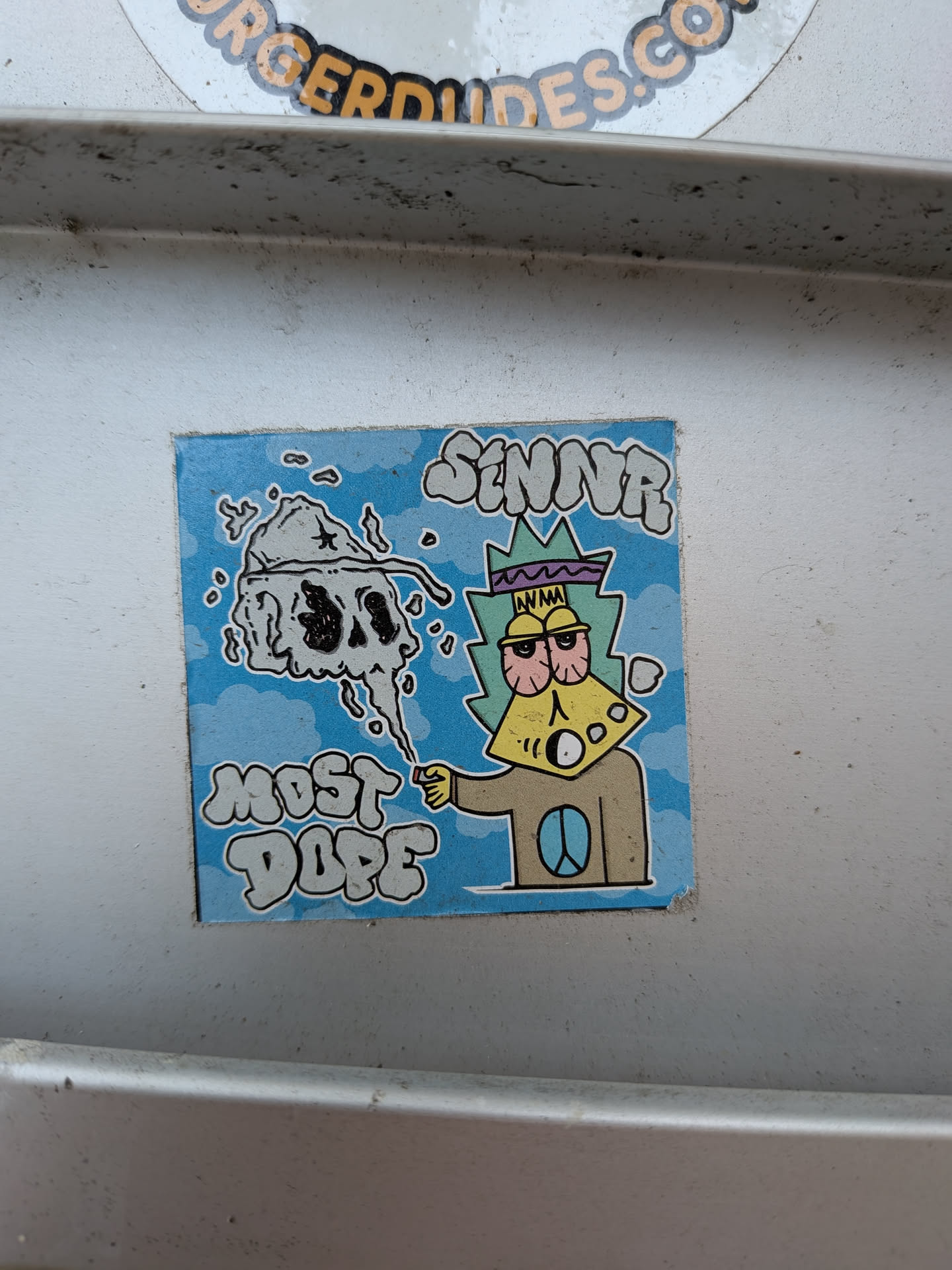 The sticker features a cartoon character with a yellow, cheese-like face, large red-rimmed eyes, and a purple and green spiky headdress. The character is wearing a brown shirt with a blue peace symbol on it. To the left of the character, a grey, smoky skull with black eyes and nose is depicted. The background is a light blue sky with white clouds. The text 'SINNR' is written in a graffiti-like font above the character's head, and 'MOST DOPE' is written in a similar font below the skull.