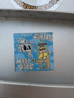 Street sticker The sticker features a cartoon character with a yellow, cheese-like face, large red-rimmed eyes, and a purple and green spiky headdress. The character is wearing a brown shirt with a blue peace symbol on it. To the left of the character, a grey, smoky skull with black eyes and nose is depicted. The background is a light blue sky with white clouds. The text 'SINNR' is written in a graffiti-like font above the character's head, and 'MOST DOPE' is written in a similar font below the skull.