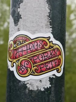 Street sticker The sticker showcases a vibrant, traditional tattoo-style design. It features a central red banner with ornate yellow and gold scrollwork and borders. The text 'TATTOOS' is prominently displayed in the upper section, with 'BY' and 'PORKY PETE' below, all rendered in a dark, stylized font. Above 'TATTOOS', a smaller curved banner reads 'EST. 1999'. The entire design is outlined in white, giving it a distinct, cut-out appearance against the dark, textured pole it's adhered to.