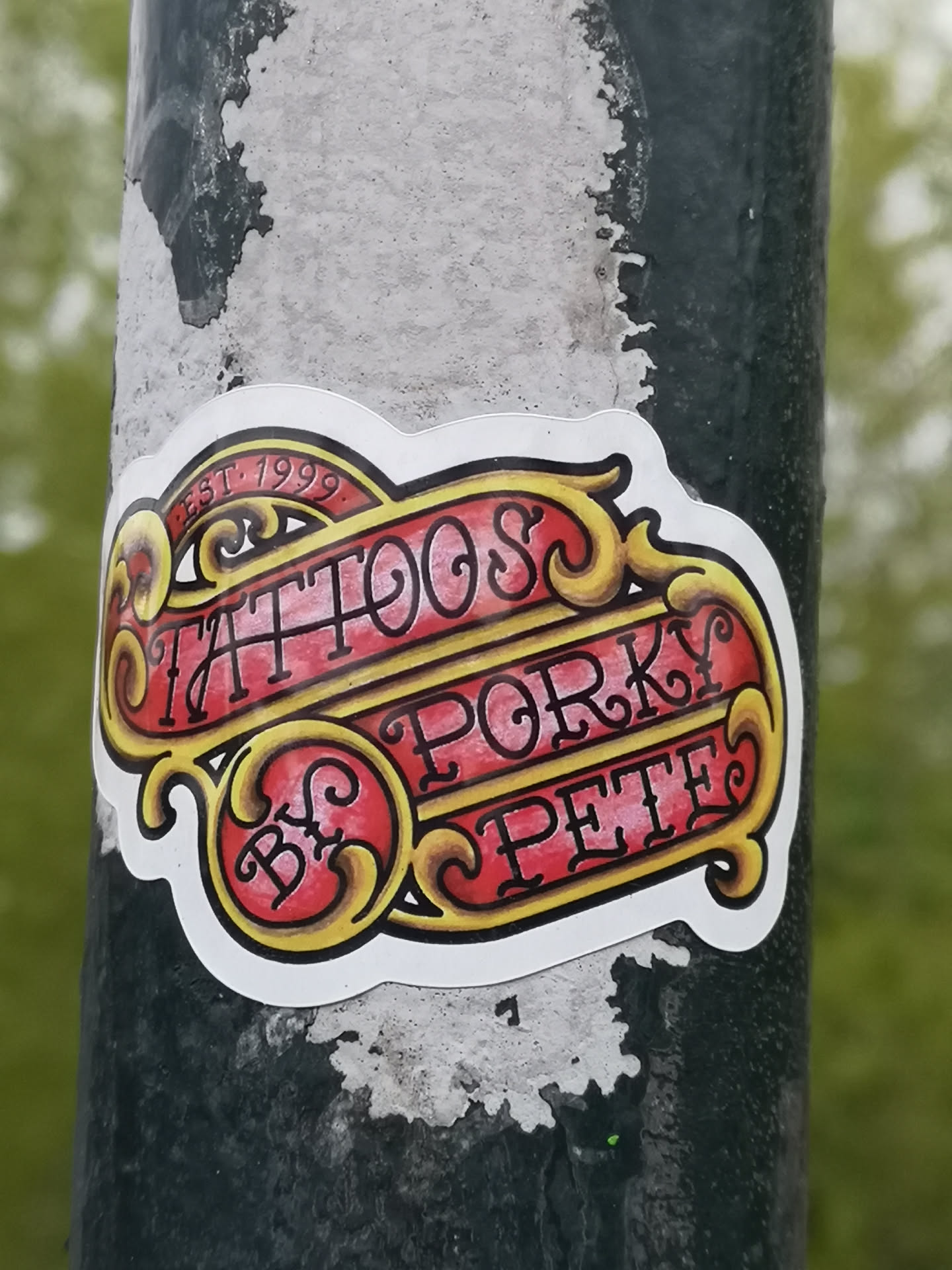 The sticker showcases a vibrant, traditional tattoo-style design. It features a central red banner with ornate yellow and gold scrollwork and borders. The text 'TATTOOS' is prominently displayed in the upper section, with 'BY' and 'PORKY PETE' below, all rendered in a dark, stylized font. Above 'TATTOOS', a smaller curved banner reads 'EST. 1999'. The entire design is outlined in white, giving it a distinct, cut-out appearance against the dark, textured pole it's adhered to.