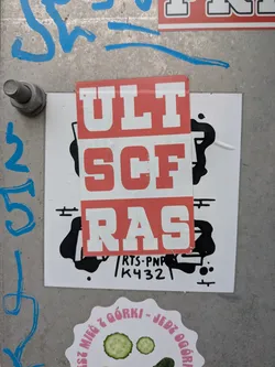 Street sticker The central sticker is a red rectangle with bold white capital letters arranged vertically to read 'ULT SCF RAS'. This sticker is placed on top of a larger white sticker that has abstract black splatter-like designs. Below the red sticker, visible on the white background, is smaller black text that says 'RTS-PNP' on one line and 'K432' on the line below it. The entire piece is affixed to a textured metal surface.