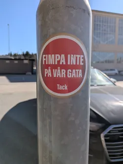 Street sticker A circular sticker with a vibrant red background, featuring a thin white inner border and a slightly wider, light brown/beige outer border. The main text, 'FIMPA INTE PÅ VÅR GATA', is prominently displayed in bold, white, sans-serif capital letters across two lines. Below this, in smaller white sans-serif letters, is the word 'Tack'. The sticker is affixed to a textured, galvanized metal pole, with some minor wear visible around its edges.