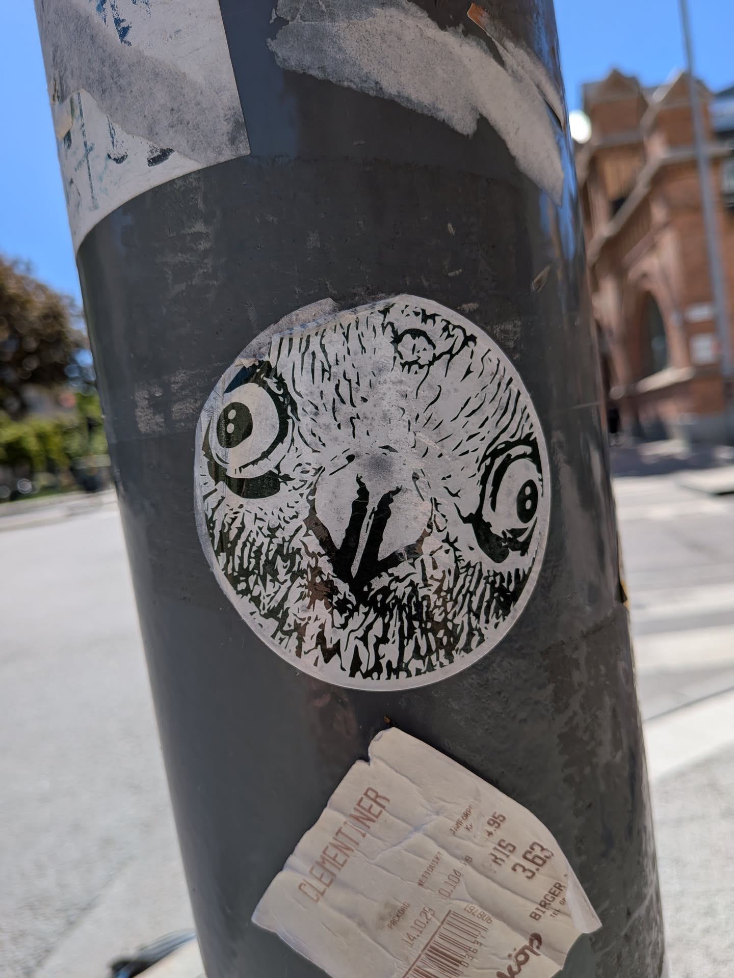 The sticker is circular, featuring a highly stylized black and white depiction of an owl's face. The design is rendered with intricate black lines and shapes on a white background, creating a textured, feathery appearance. The owl has very large, prominent round eyes with small, dark pupils, giving it an intense or surprised expression. A distinct V-shaped beak is visible in the center, below the eyes. The overall aesthetic is graphic and somewhat abstract, focusing on the owl's facial features.