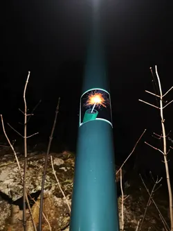 Street sticker The sticker features a stylized illustration of a lit firecracker or dynamite stick. It is rectangular with slightly rounded corners, framed by a thin white border. The internal background of the sticker is dark, almost black, creating a strong contrast for the main subject. The firecracker itself is depicted in a bright green color, with a white fuse extending upwards. The fuse culminates in a brilliant, starburst-like sparkler effect, radiating intense orange, yellow, and white light, simulating an active ignition.