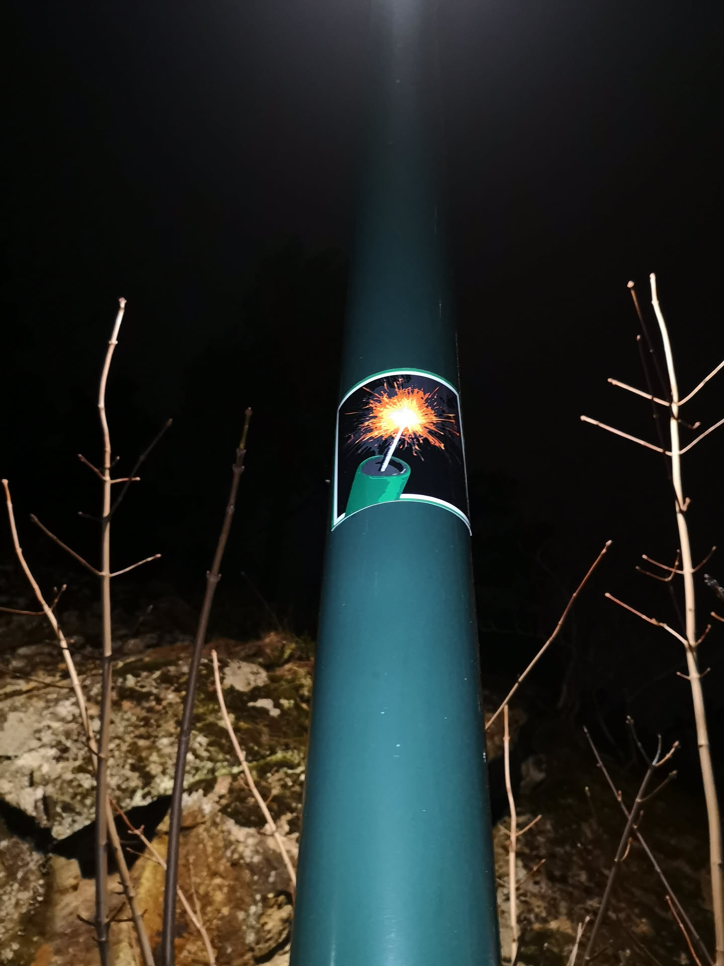 The sticker features a stylized illustration of a lit firecracker or dynamite stick. It is rectangular with slightly rounded corners, framed by a thin white border. The internal background of the sticker is dark, almost black, creating a strong contrast for the main subject. The firecracker itself is depicted in a bright green color, with a white fuse extending upwards. The fuse culminates in a brilliant, starburst-like sparkler effect, radiating intense orange, yellow, and white light, simulating an active ignition.