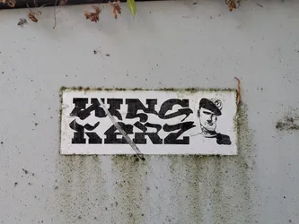 A rectangular white sticker with black print. Features bold, stylized graffiti text reading 'WINS KERZ' alongside a line-art portrait of a man in a cap with neck tattoos. The sticker is weathered with a diagonal scratch and mossy edges.