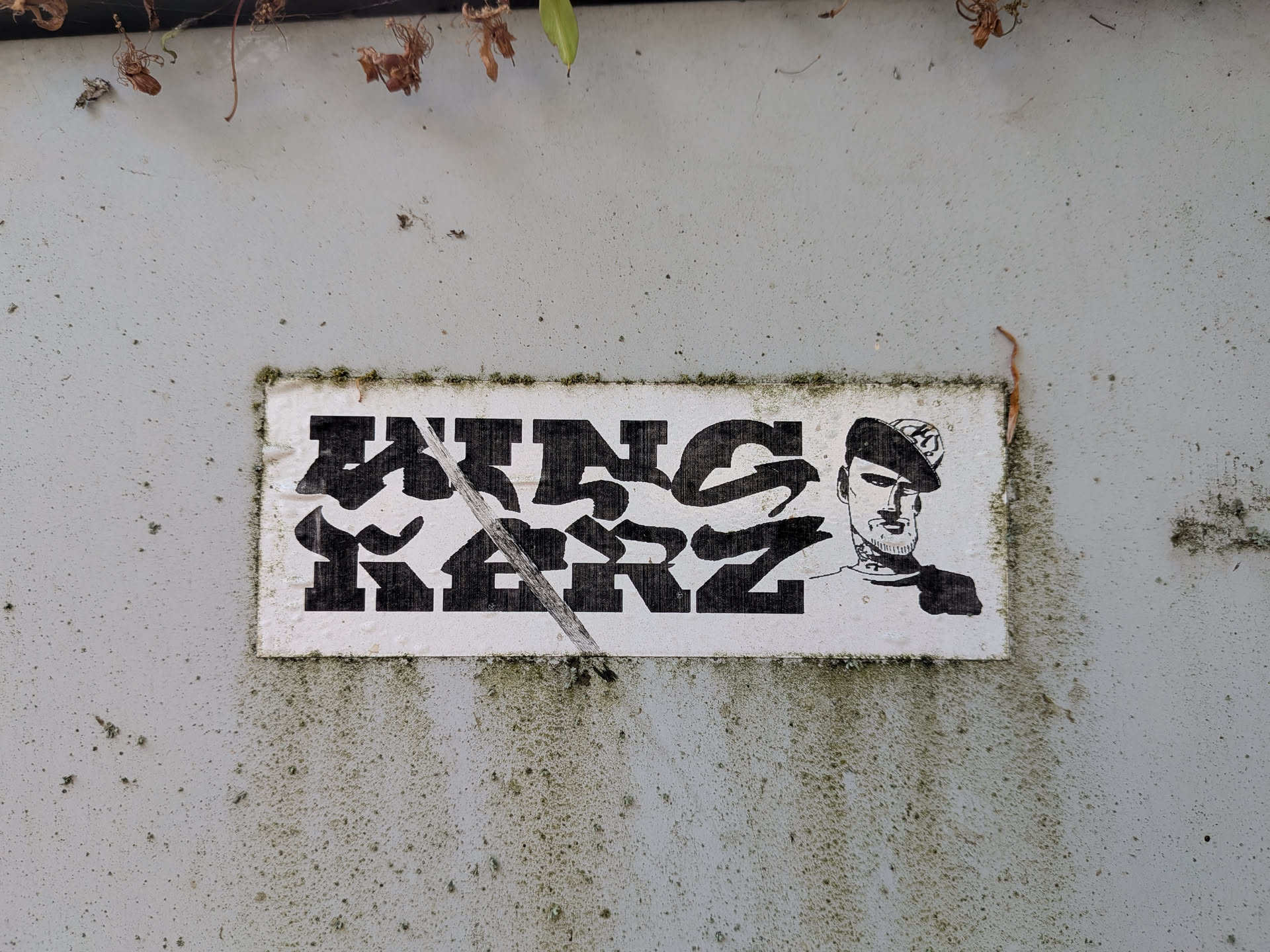 A rectangular white sticker with black print. Features bold, stylized graffiti text reading 'WINS KERZ' alongside a line-art portrait of a man in a cap with neck tattoos. The sticker is weathered with a diagonal scratch and mossy edges.