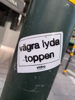 A sticker on a pole with the text "Vägra lyda toppen" (Refuse to obey the top) and the word "vidro" at the bottom. The text is in a bold, sans-serif font.  The sticker is white with black text.
