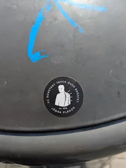 Street sticker The sticker is black and circular, with white text and a white graphic. The central graphic is a minimalist silhouette of a human torso viewed from the back, highlighting the spine. Protruding from the spine are multiple short, sharp objects resembling needles or pins. Around the top curve of the circle, the text "FC KONSTANT TRYCK ÖVER BRÖSTET" is written in white capital letters. Around the bottom curve, the text "JÄMNA PLÅGOR" is visible, also in white capital letters. Below the central figure, a smaller text 'Est. 2025' is present. The design has a somber or unsettling tone, associating the imagery with the textual phrases.